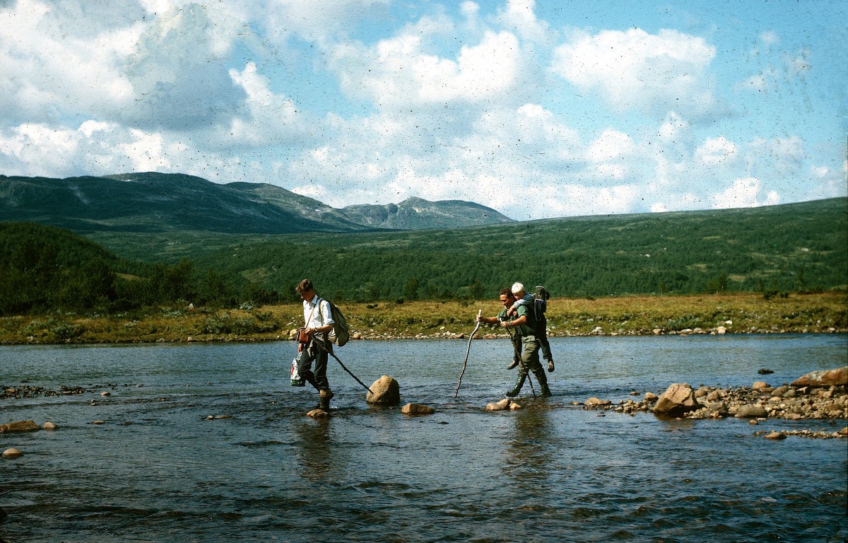 To personer krysser en elv i norsk fjellandskap, med tursekker og staver. Klar himmel og fjell i bakgrunnen.