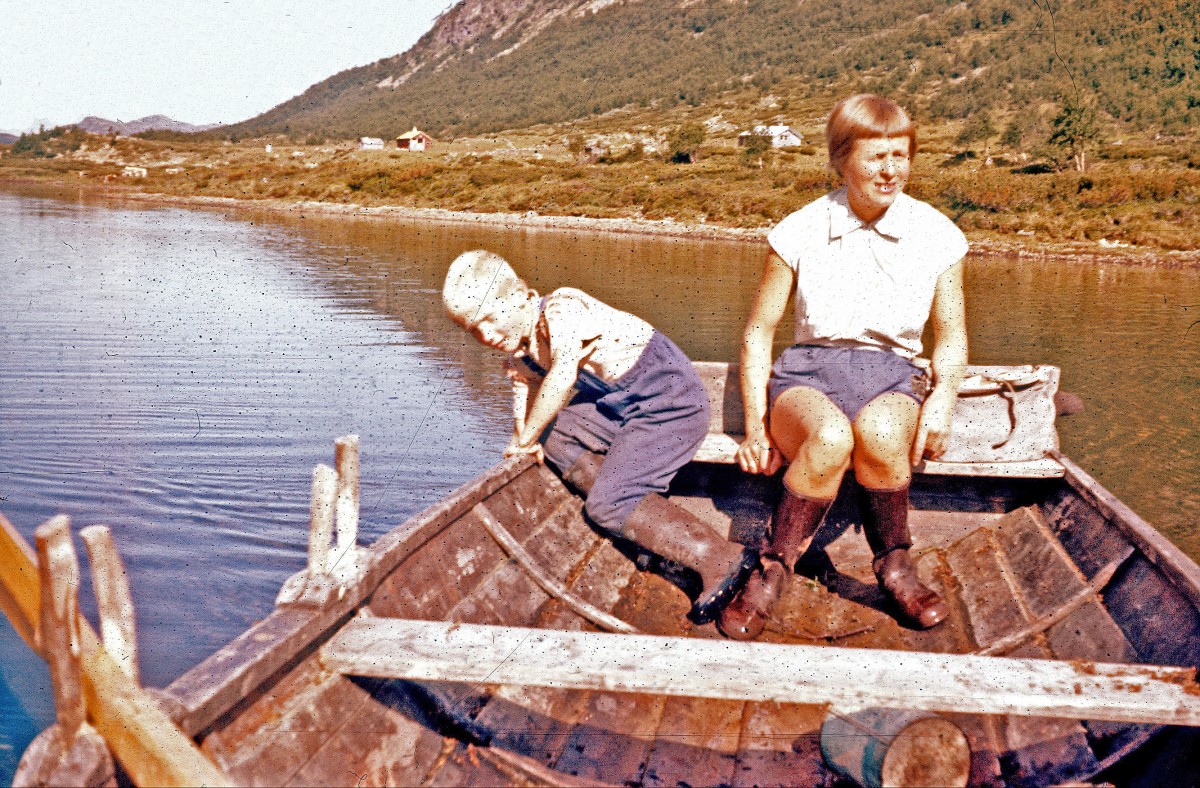 To personer sitter i en trebåt på en innsjø med fjell og hytte i bakgrunnen.