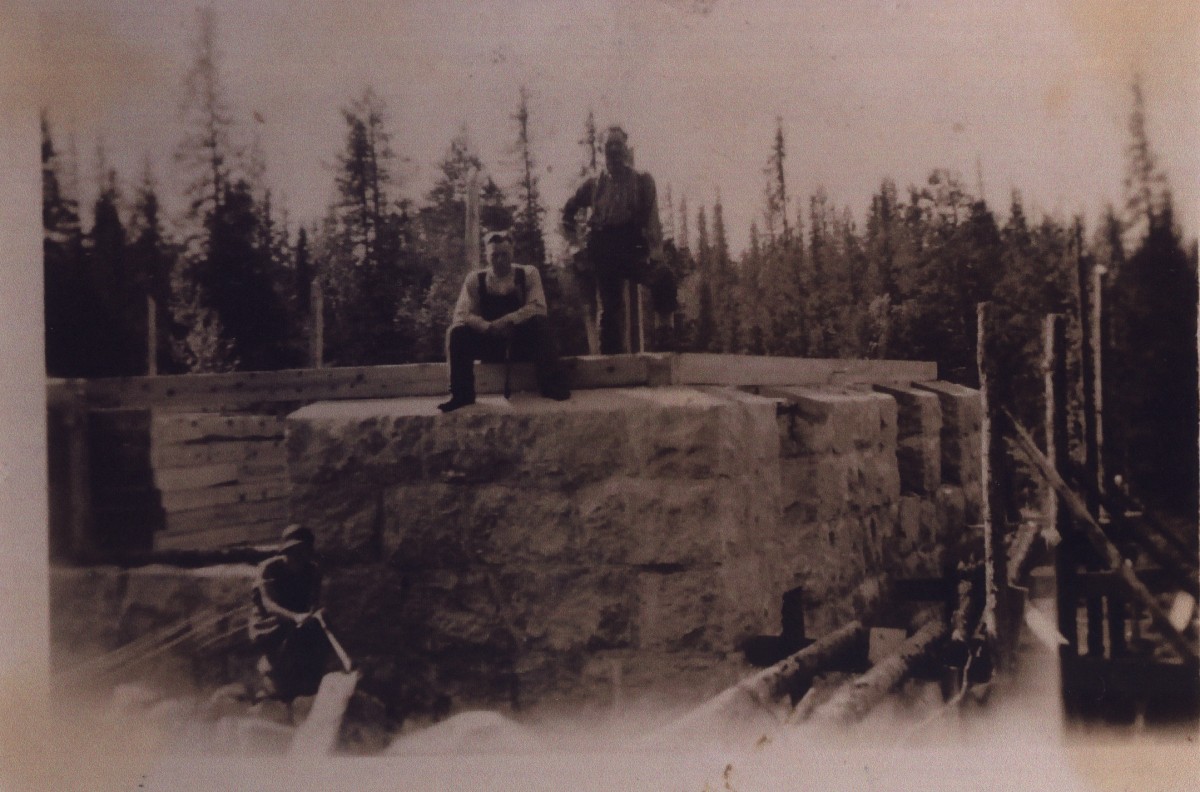 Svart-hvitt bilde av tre menn som jobber med steinbygning i skogsområde.