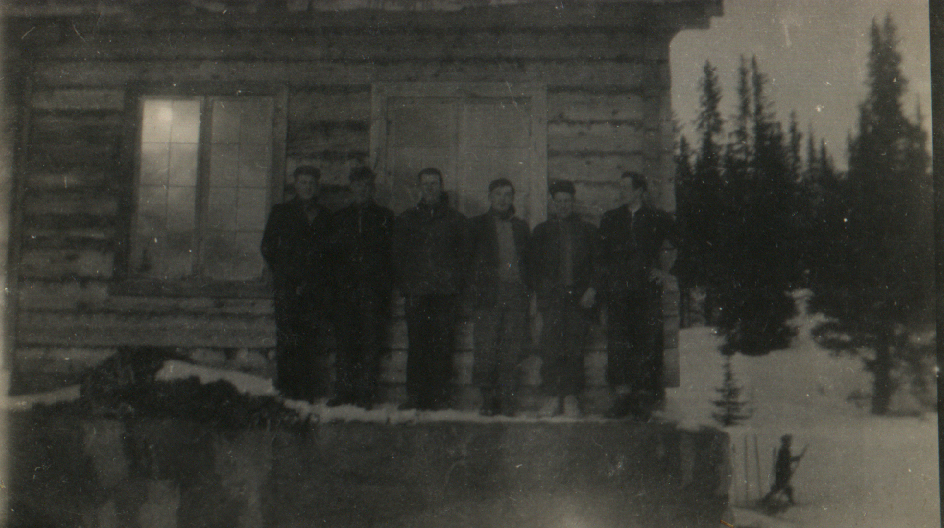 Gruppe menn foran en hytte i snødekt skoglandskap, natt.Vintage sort-hvitt bilde, tåkete og mystisk stemning.