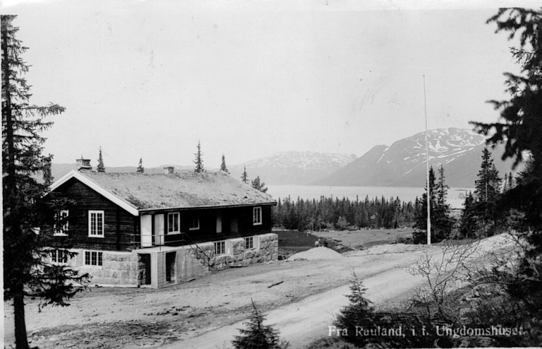 Eldre hytte i norsk fjellandskap med skog og snødekte fjell i bakgrunnen.