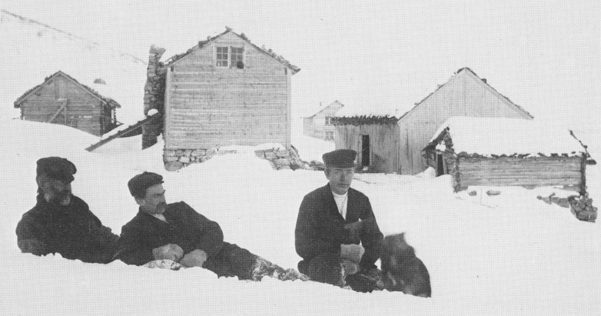 Svart kvit bilde. Vinterlandskap med gard i bakgrunnen, tre mennog ein hund framme i bildet