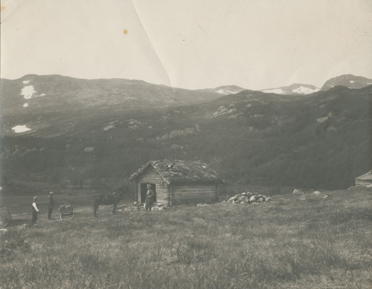 Gammel hytte i norsk fjellandskap med hest og folk i forgrunnen. Snøkledde fjell i bakgrunnen.