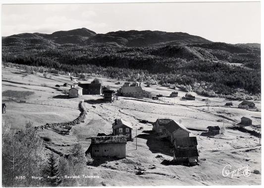 Bygdelandskap i Rauland, Telemark, med fjell i bakgrunnen. Svart-hvitt foto med gårdsbygninger og åser.