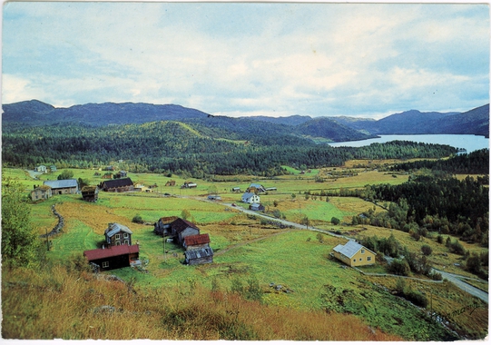 Landskap i Norge med grønne åser, tradisjonelle trehus og en innsjø i bakgrunnen på en klar dag.