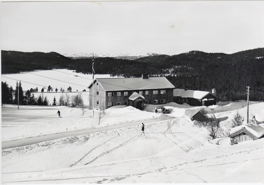 Snødekt landskap med skikjørere og stor hytte ved skogen en vinterdag.