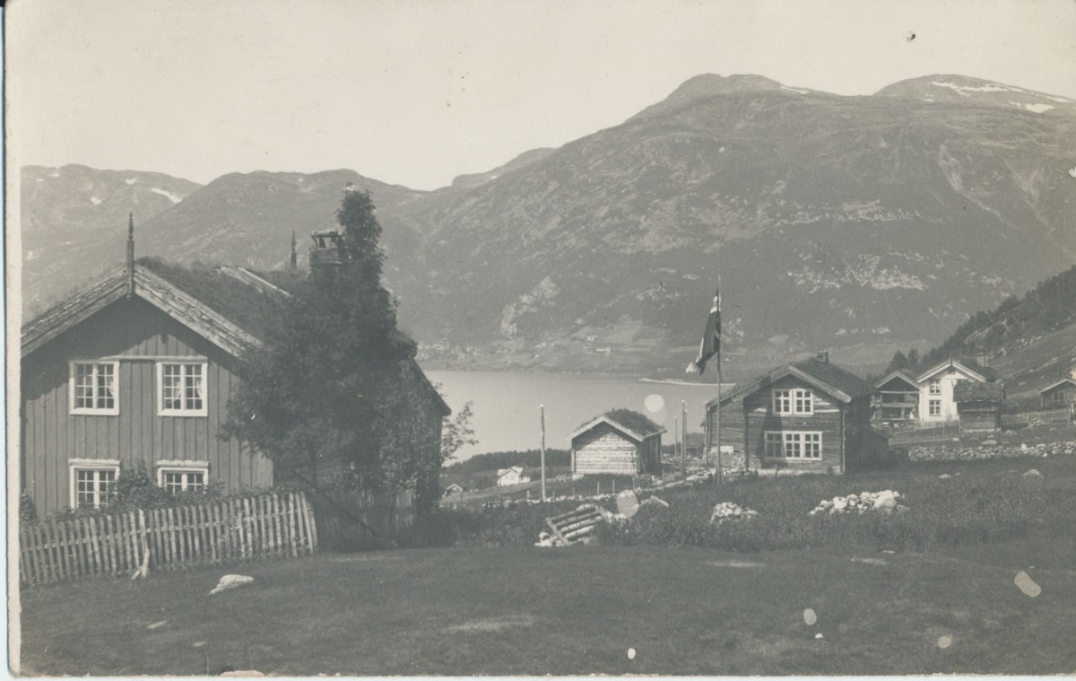 Gammelt norsk landskap med trehus, fjord og fjell i bakgrunnen, fanget i et nostalgisk sort-hvitt bilde.