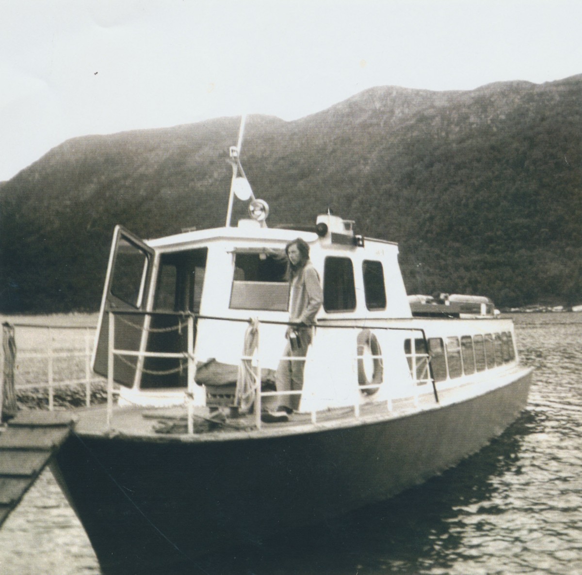 Vintage bilde av en person på en båt ved kai, omgitt av fjellandskap og vann.