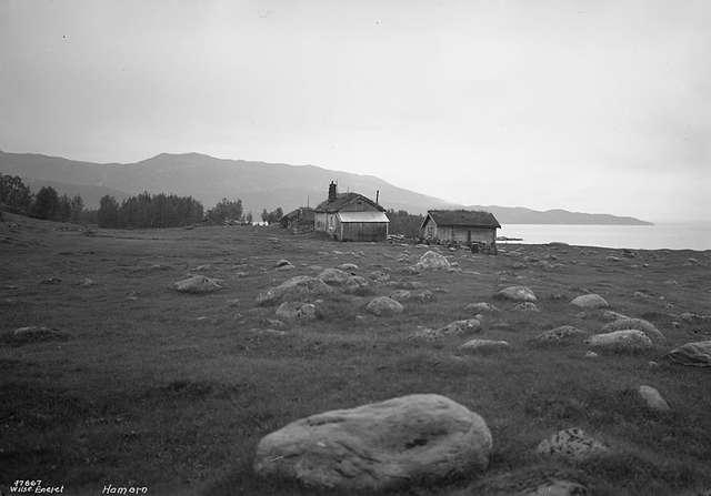 Gammel norsk gård på landsbygda med gressletter, fjell i bakgrunnen og bebyggelse ved havet, Hamarøy.