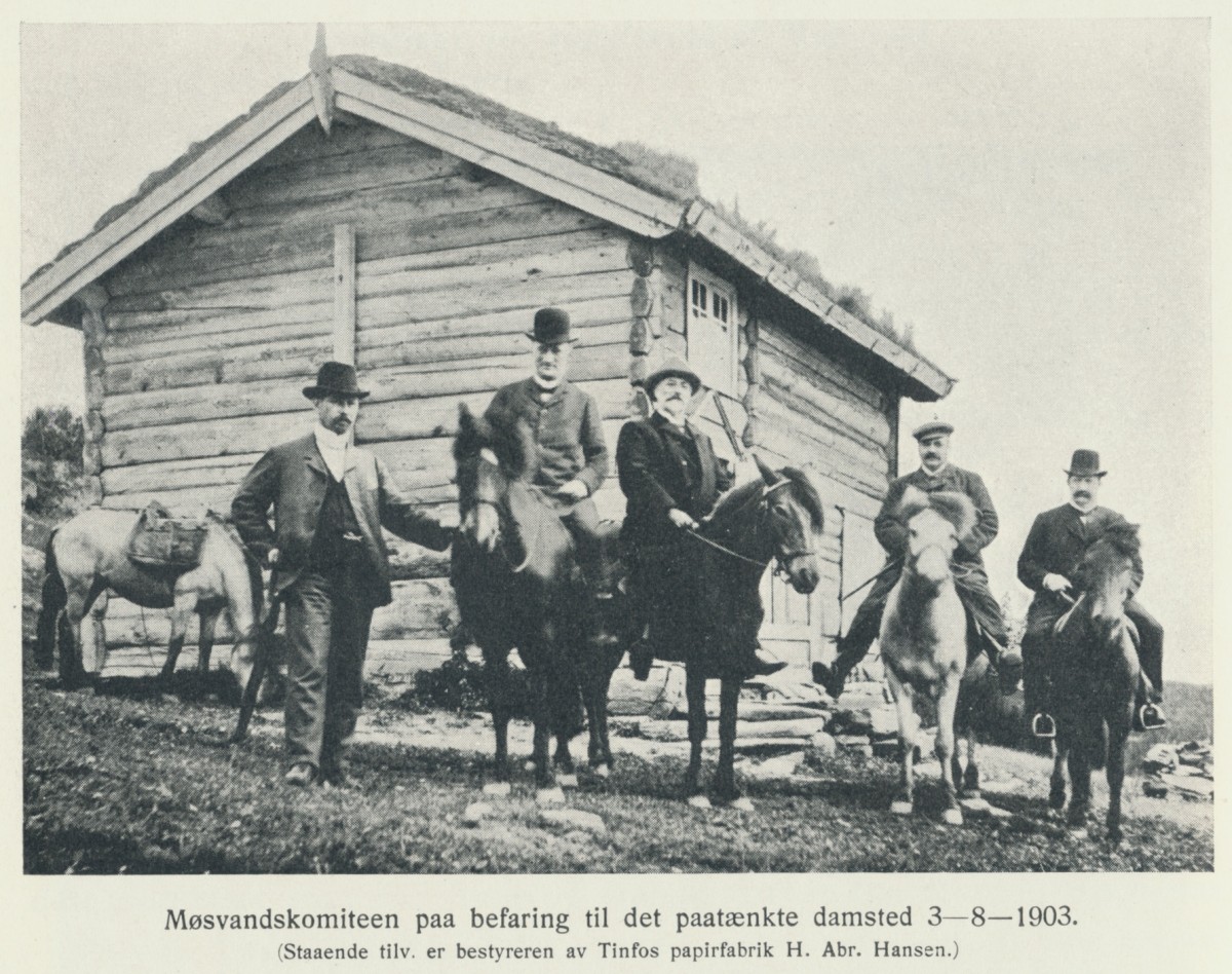 Menn foran en tømmerhytte med hester i 1903, relatert til Møsvandskomiteens befaring til damstedet.