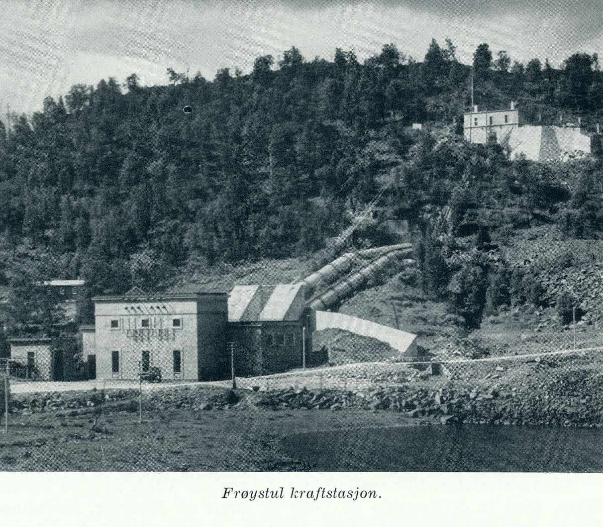 Kraftstasjon på Frogstul omringet av skogklædte fjell, med rørledninger og bygninger i forgrunnen.