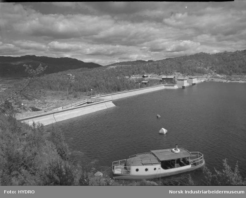 Svart-hvitt bilde av en dam i Norge med båt i forgrunnen, omgitt av skog og fjelllandskap, overskyet himmel.