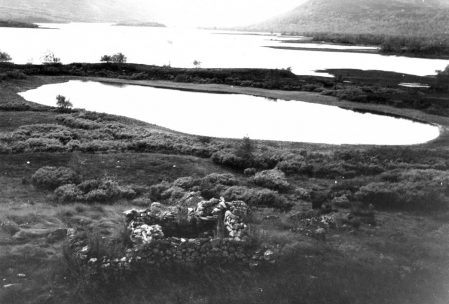 Svart-hvitt landskapsbilde av innsjø med fjell i bakgrunnen, omkranset av frodig vegetasjon og steinformasjoner.