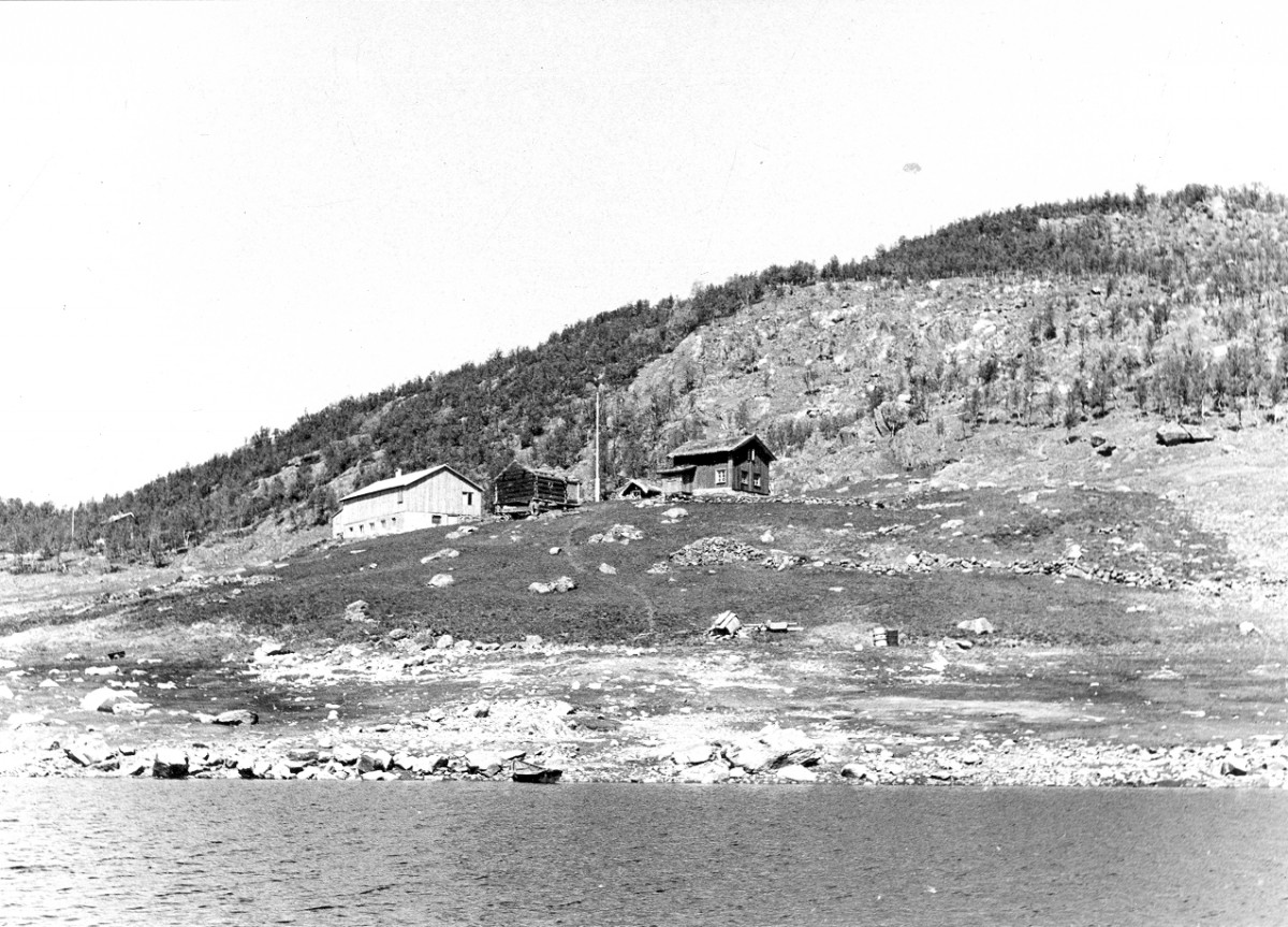 Svarthvitt bilde av trehytter ved en innsjø, omgitt av fjellandskap.