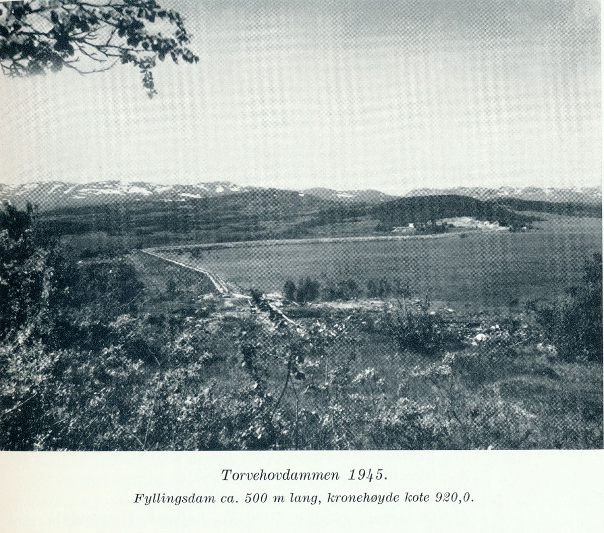 Svart-hvitt bilde av Torvehovdammen i 1945, fjellandskap i bakgrunnen, fyllingsdam ca. 500 m lang.