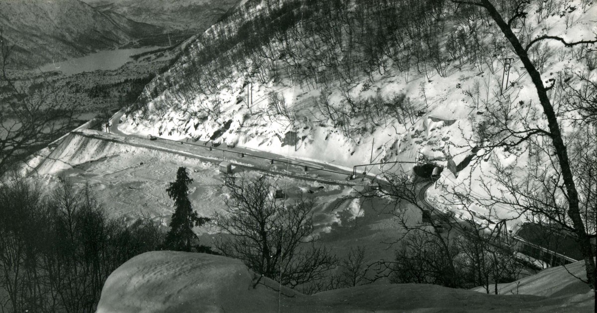 Snødekt vei gjennom fjellandskap i vinteren, med innsjø i bakgrunnen.