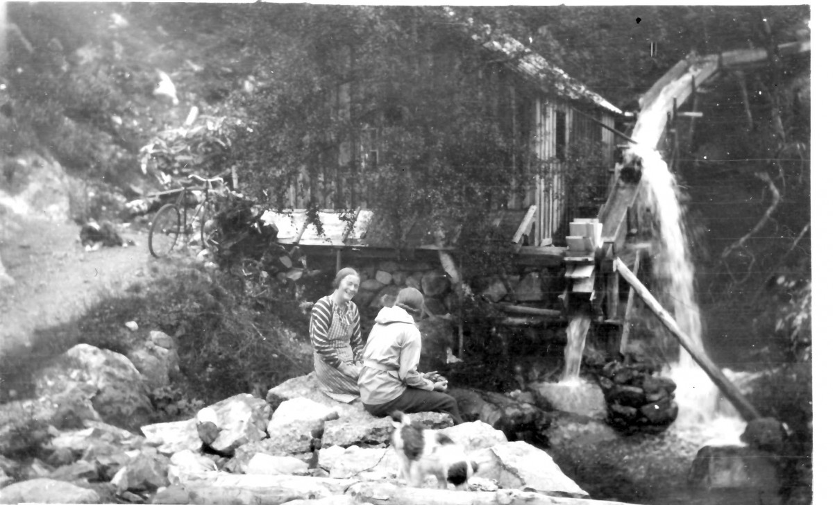 To kvinner ved en gammel vannmølle i skogen, med en sykkel i bakgrunnen og en hund på steinene foran.