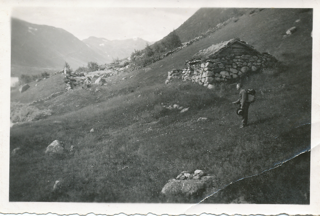 Gammel hytte i norsk fjellandskap med person som går i gresset. Svart-hvitt bilde med fjell utsikt.
