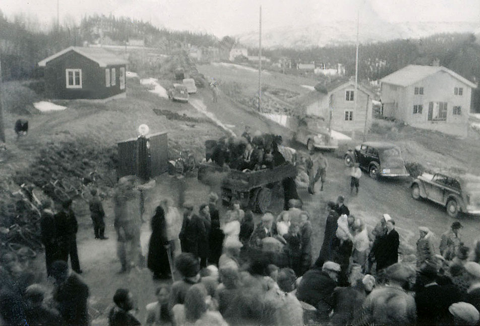 Sort-hvitt bilde av en folkemengde og biler i en liten norsk bygd på 1940-tallet.