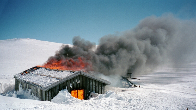 Brann i en snødekket hytte med tykk røyk mot blå himmel. Flammer synlige i det kalde, hvite vinterlandskapet.
