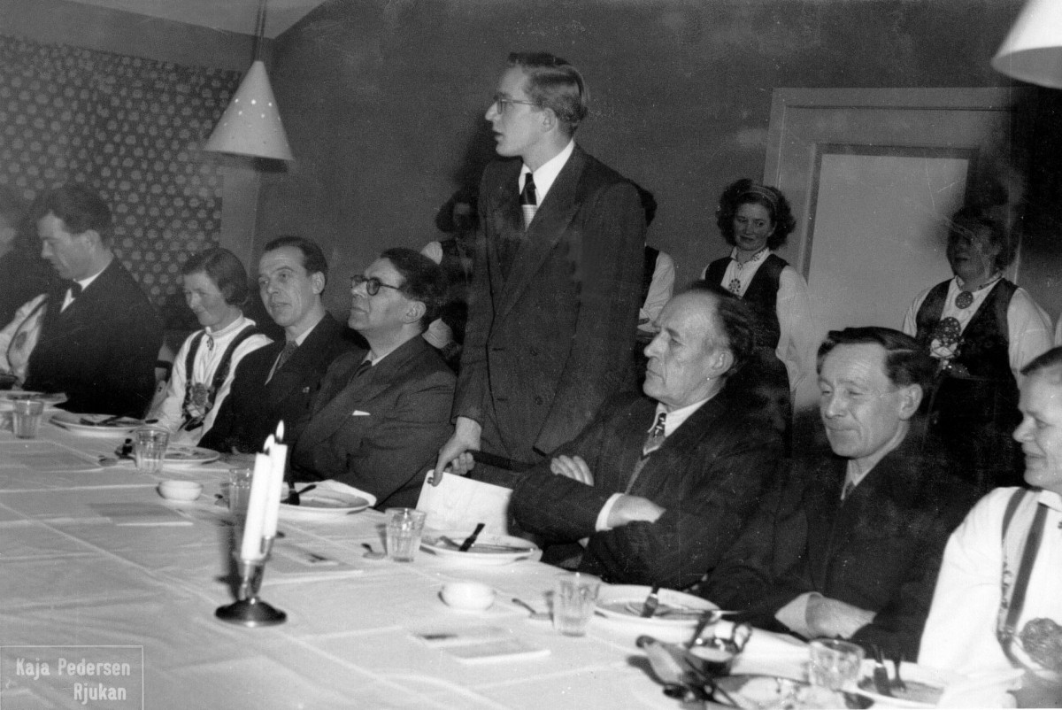 Gruppe mennesker i formelt antrekk rundt et bord under et arrangement, med en mann stående og snakkende.