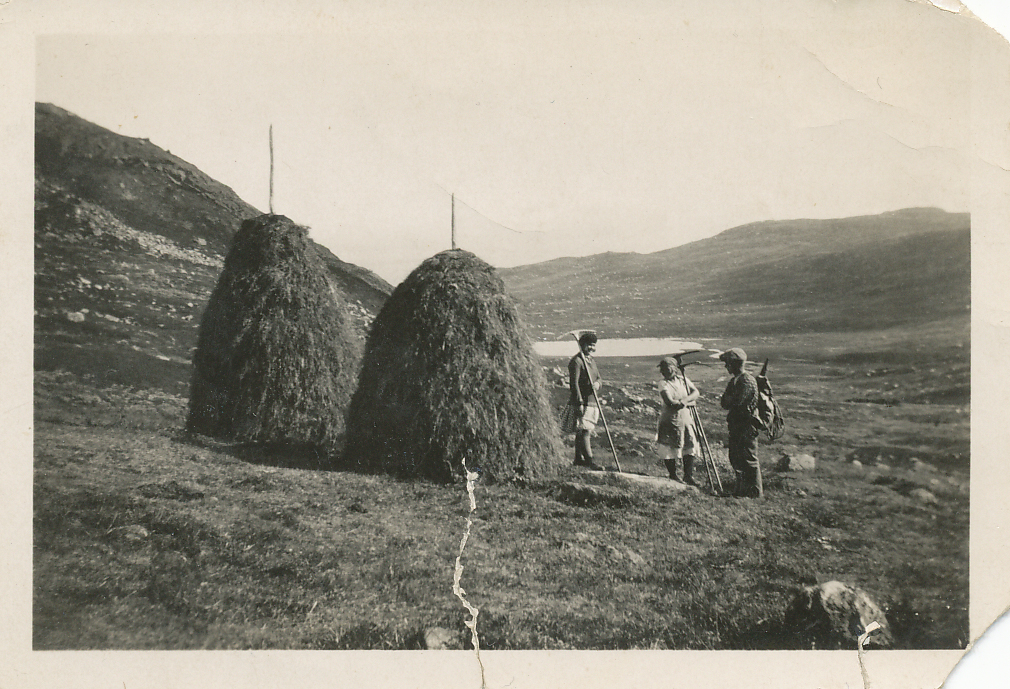 Tre vandrere i fjellandskap nær to høystakker. Utsikt over dal og innsjø i bakgrunnen. Vintage foto i svart-hvitt.