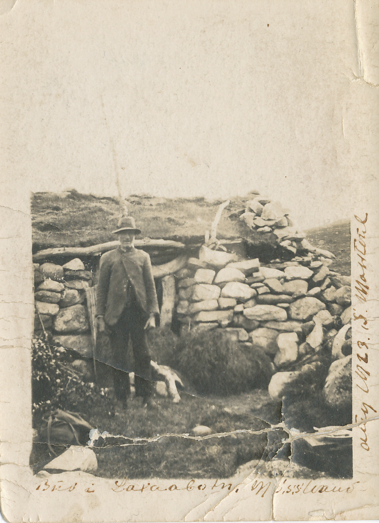 Gammel fotografi av mann foran steinhytte i tradisjonelle klær. Historisk bilde med landlig atmosfære.