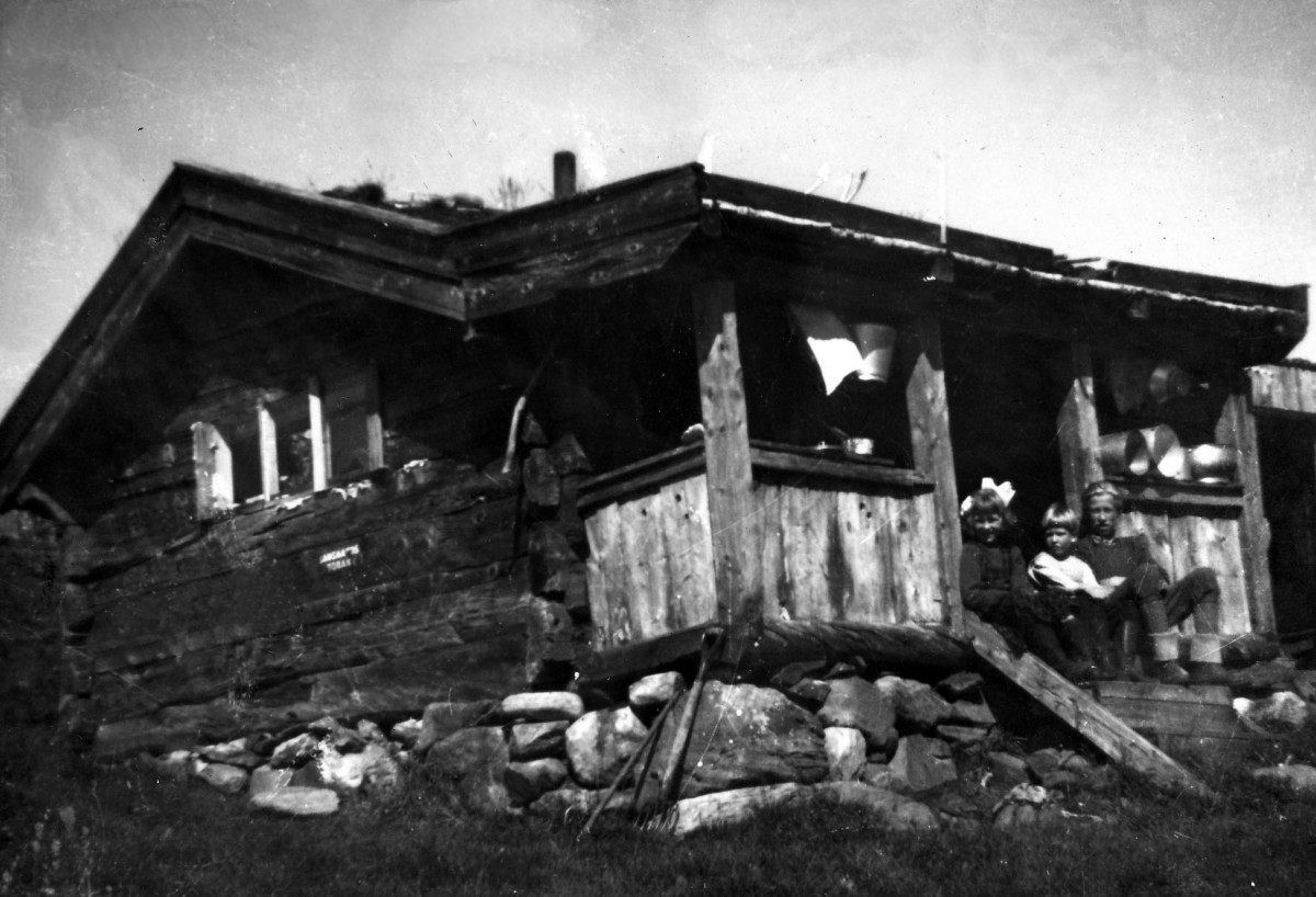 Gamle trehus med barn på trappen, omgitt av natur og steiner, nostalgisk svart-hvitt foto.