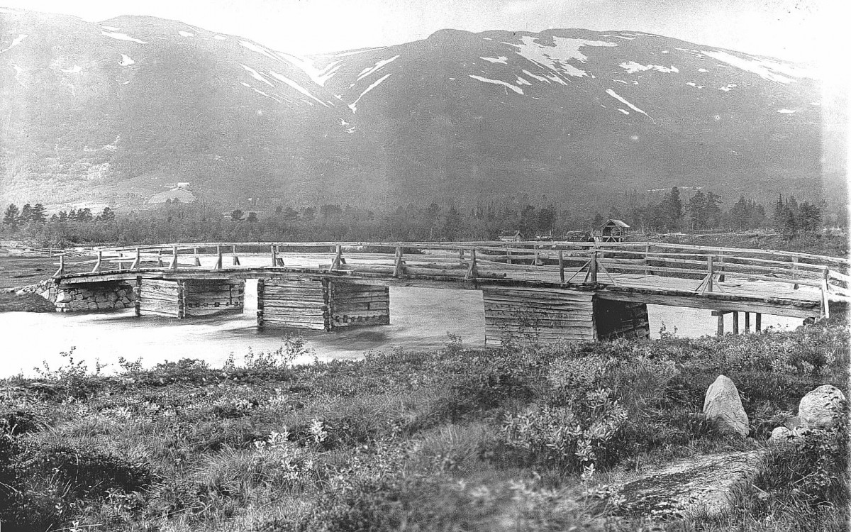 Gamle trebro over elv med snødekte fjell i bakgrunnen. Naturskjønt landskap i sort-hvitt bilde.