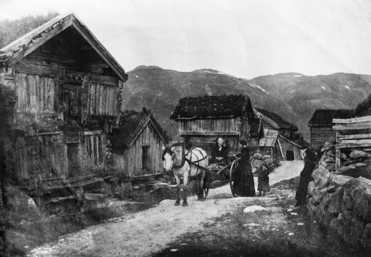 Gammelt norsk gårdstun med personer, hest og kjerre, omgitt av fjell og tradisjonelle tømmerhus.