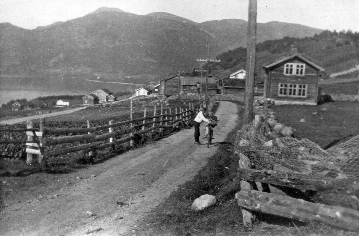 Landlig vei i Norge med syklist, omgitt av gamle trehus og fjell i bakgrunnen. Svart-hvitt foto.