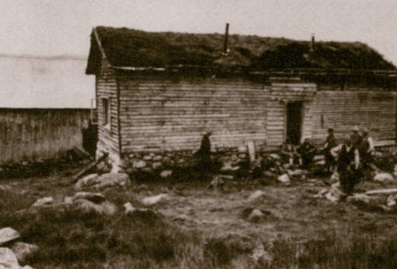 Gammelt trehus i et landlig landskap, delvis dekket av gress. Flere personer står ved siden av bygningen.