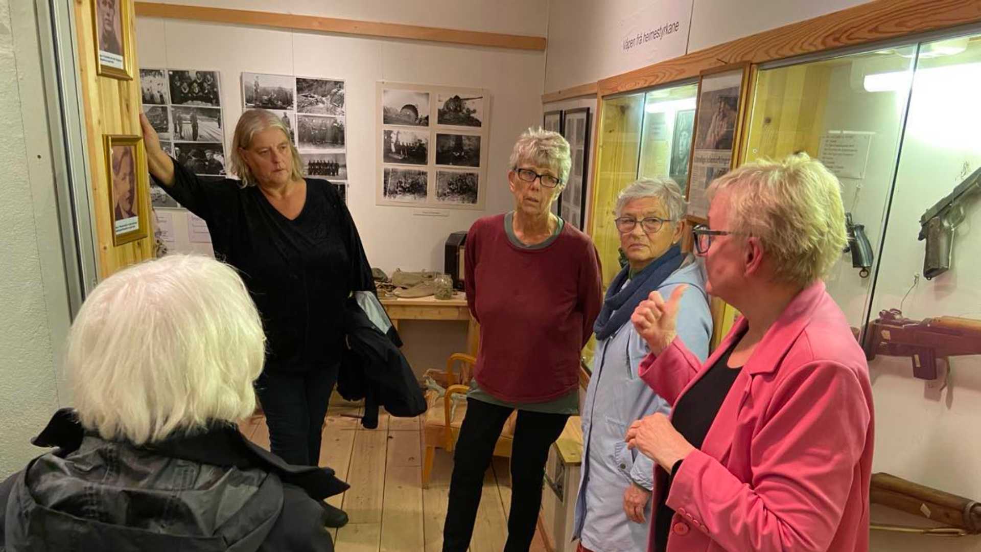 Gruppe med kvinner diskuterer utstilling i museum; historiske fotografier og våpen vises.