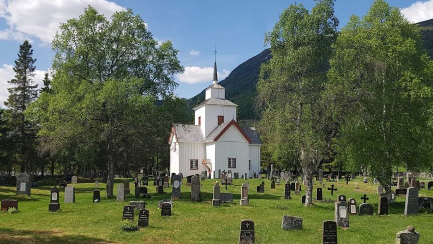 Hvit, historisk kirke omringet av gravstøtter og grønne trær, med blå himmel og fjell i bakgrunnen.