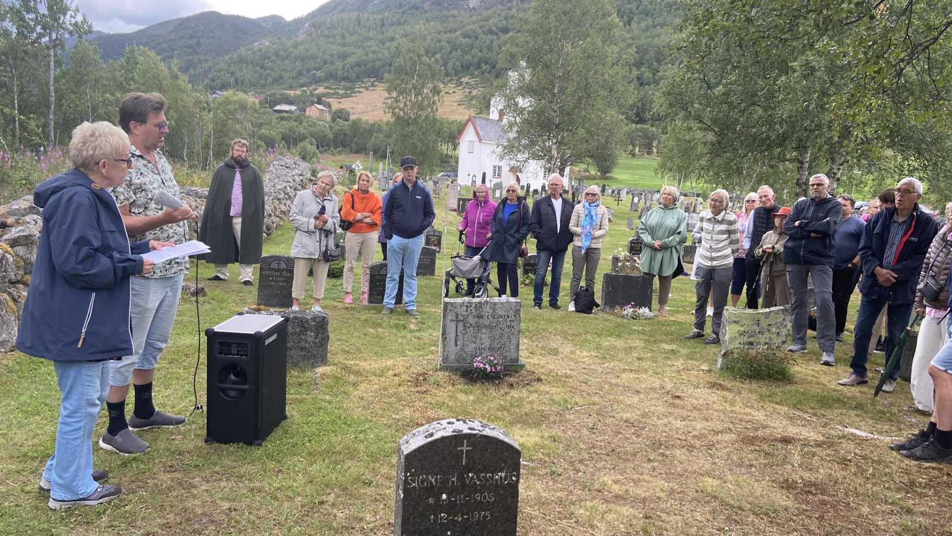 Gruppe mennesker samlet ved en gravplass i naturskjønne omgivelser, lytter til en taler.