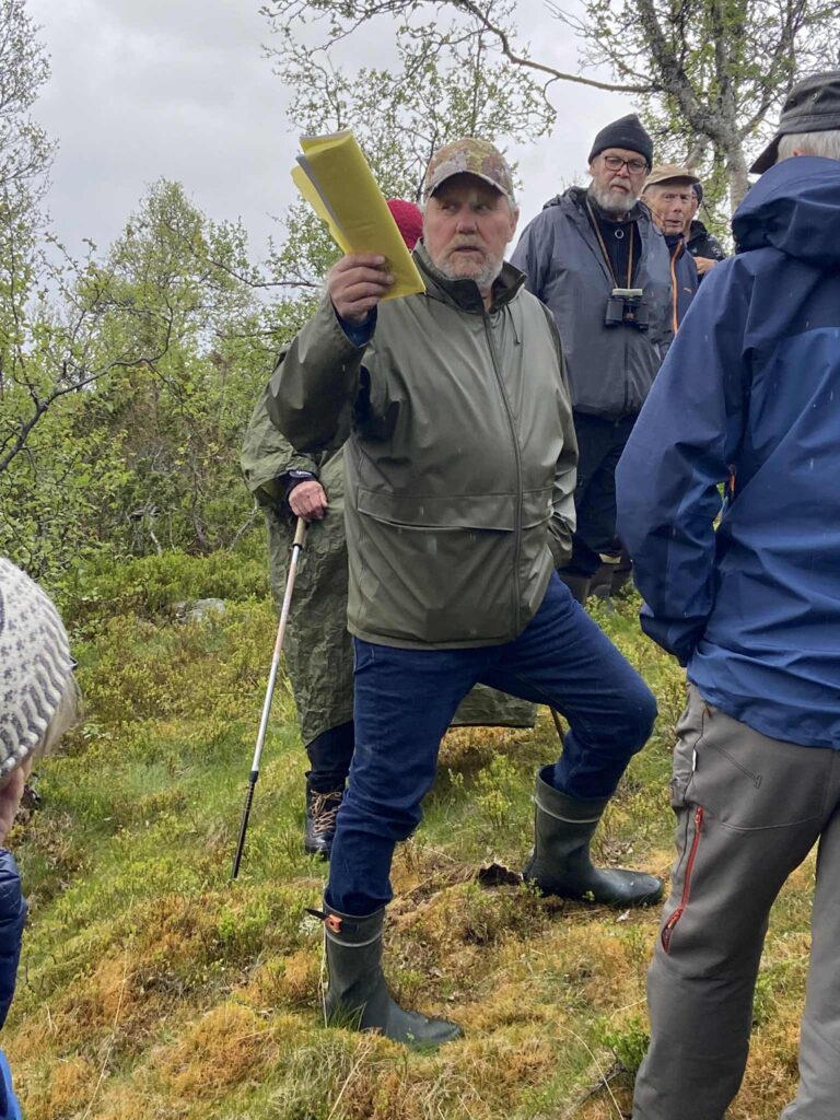 En mann holder et papir og leder en tur i skogen, omgitt av andre voksne iført turklær.