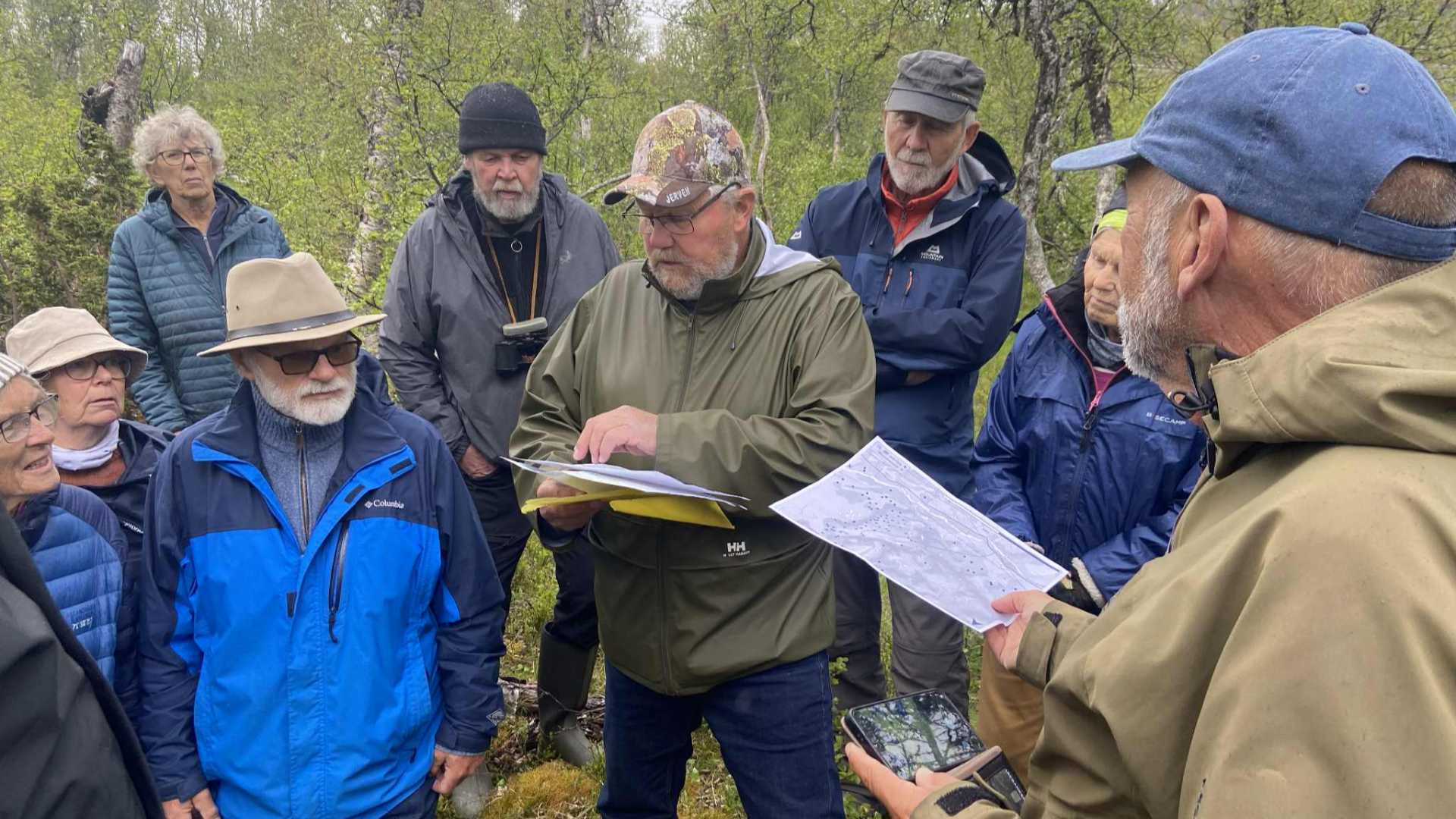 Mennesker samles i skogen rundt en mann som viser kart og forklarer, trolig på en guidet naturopplevelse.