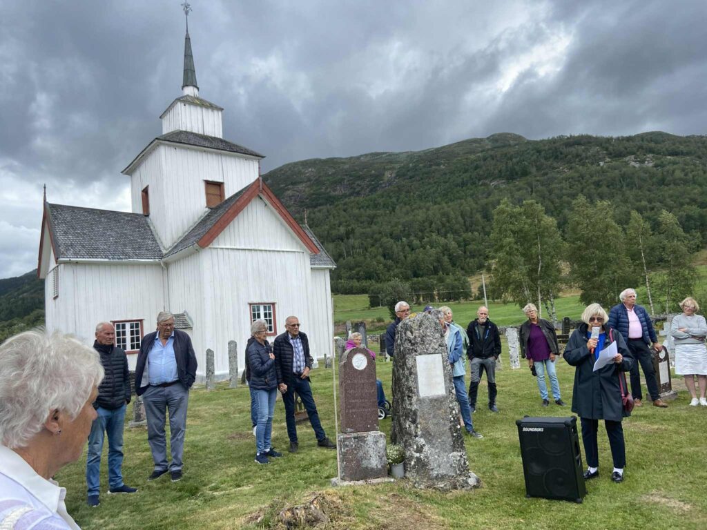 Gruppe foran hvit kirke, samling i vakker natur, dramatisk skydekket.