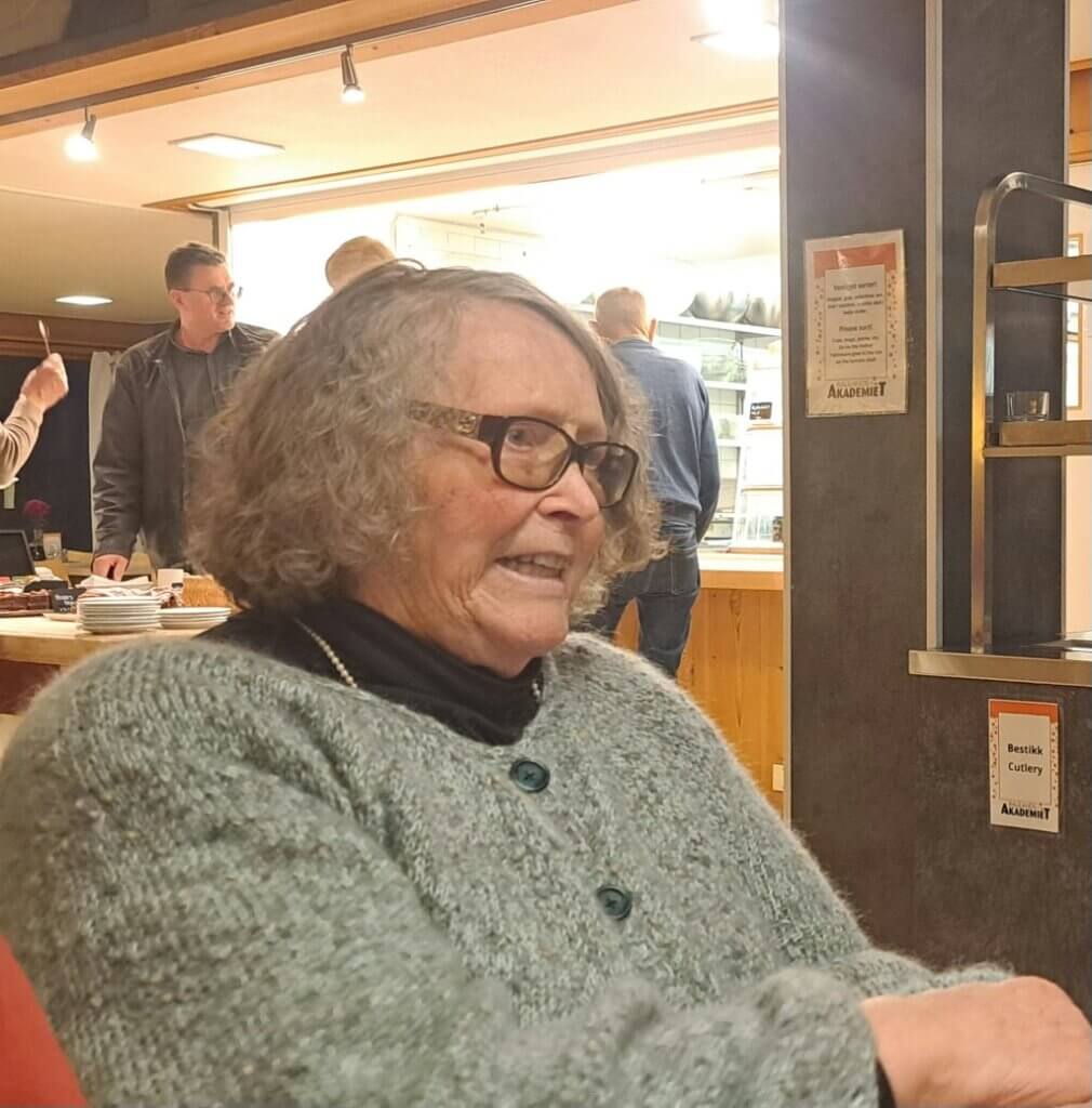Eldre kvinne med briller smiler innendørs, med mennesker i bakgrunnen.