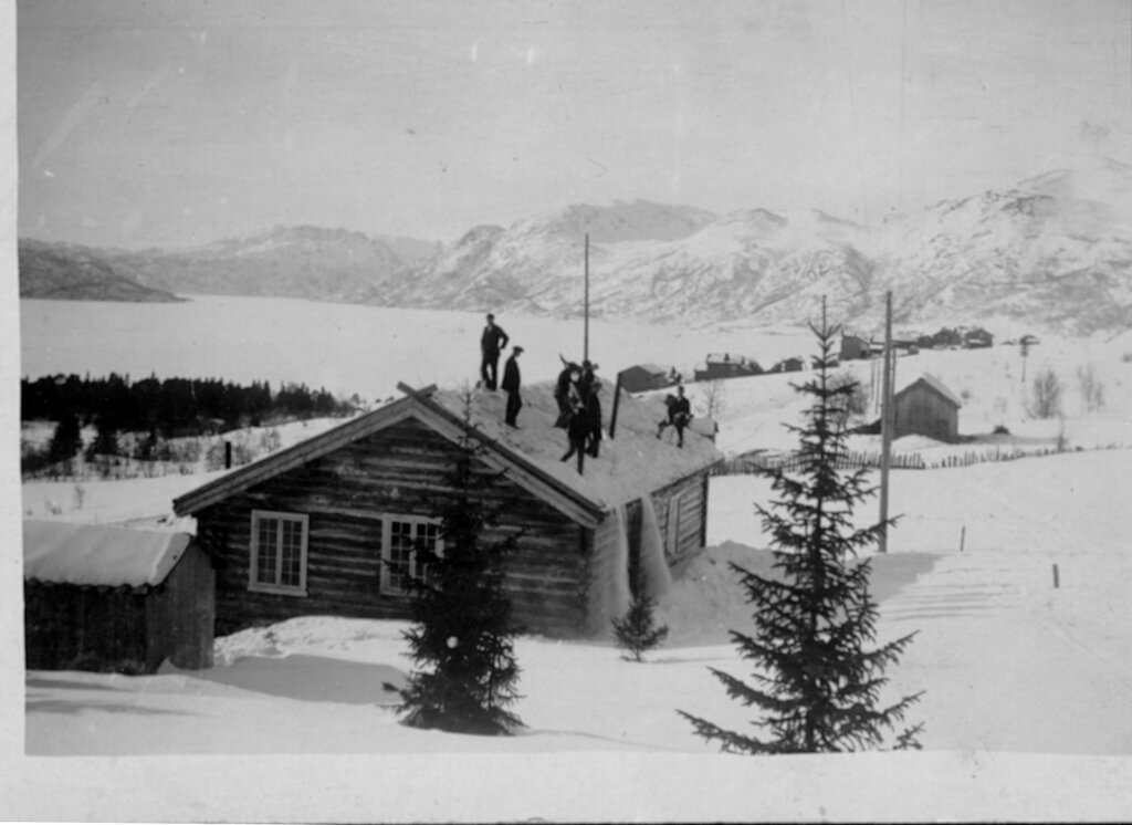 Personer på snødekt tak i norsk vinterlandskap, omgitt av fjell og trær.