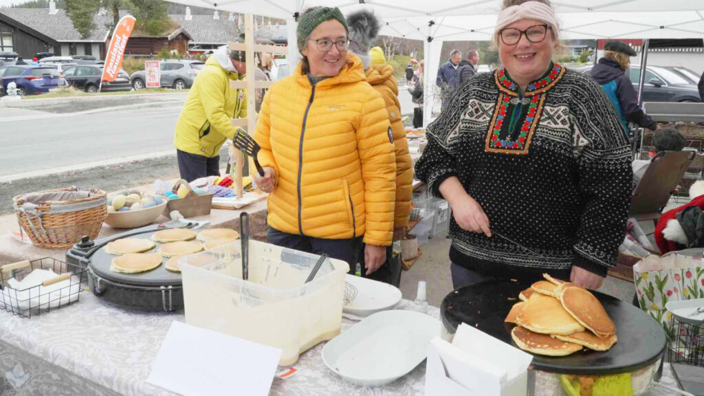 To kvinner steker pannekaker på marked, iført fargerike klær. Utsalg under åpent telt med flere besøkende.