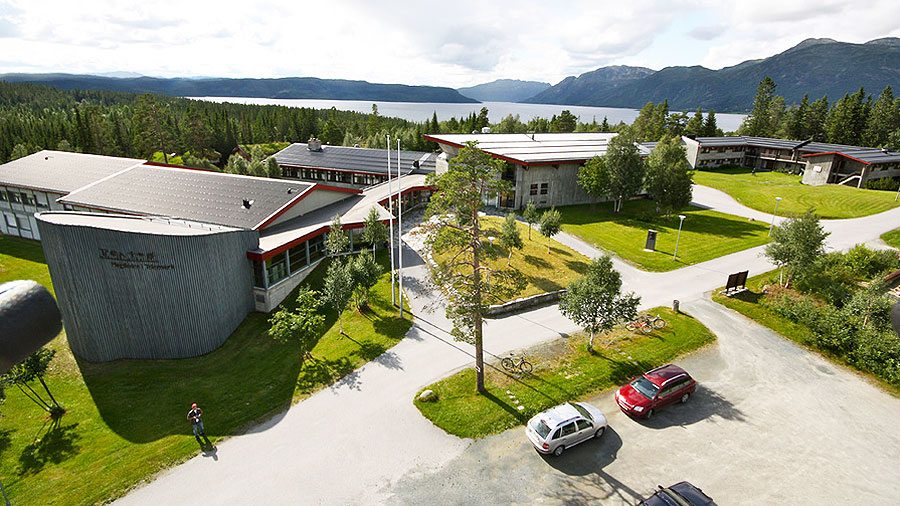 Utsikt over et moderne skolekompleks omgitt av natur, med fjell og vann i bakgrunnen, på en solfylt dag.