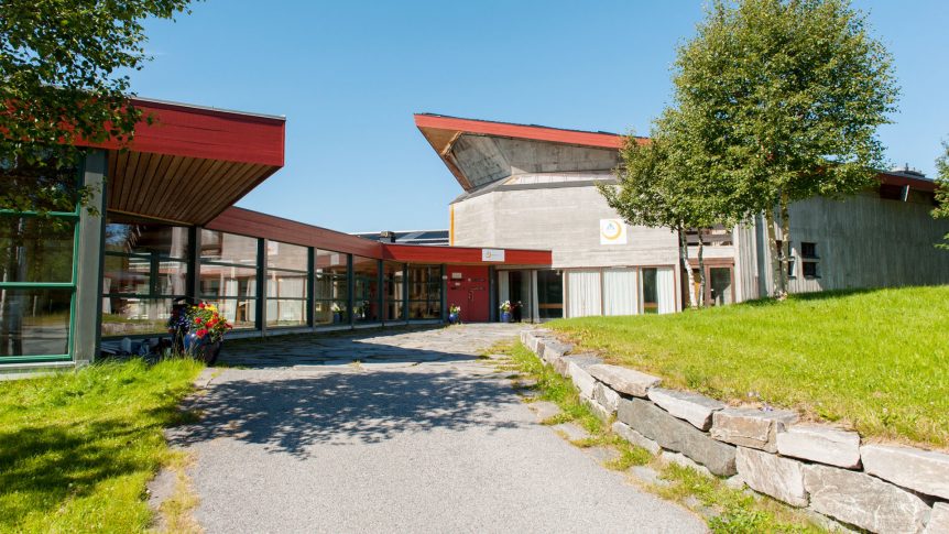 Moderne bygning med rødt tak og glassfasade, omgitt av grønt gress og trær under blå himmel.