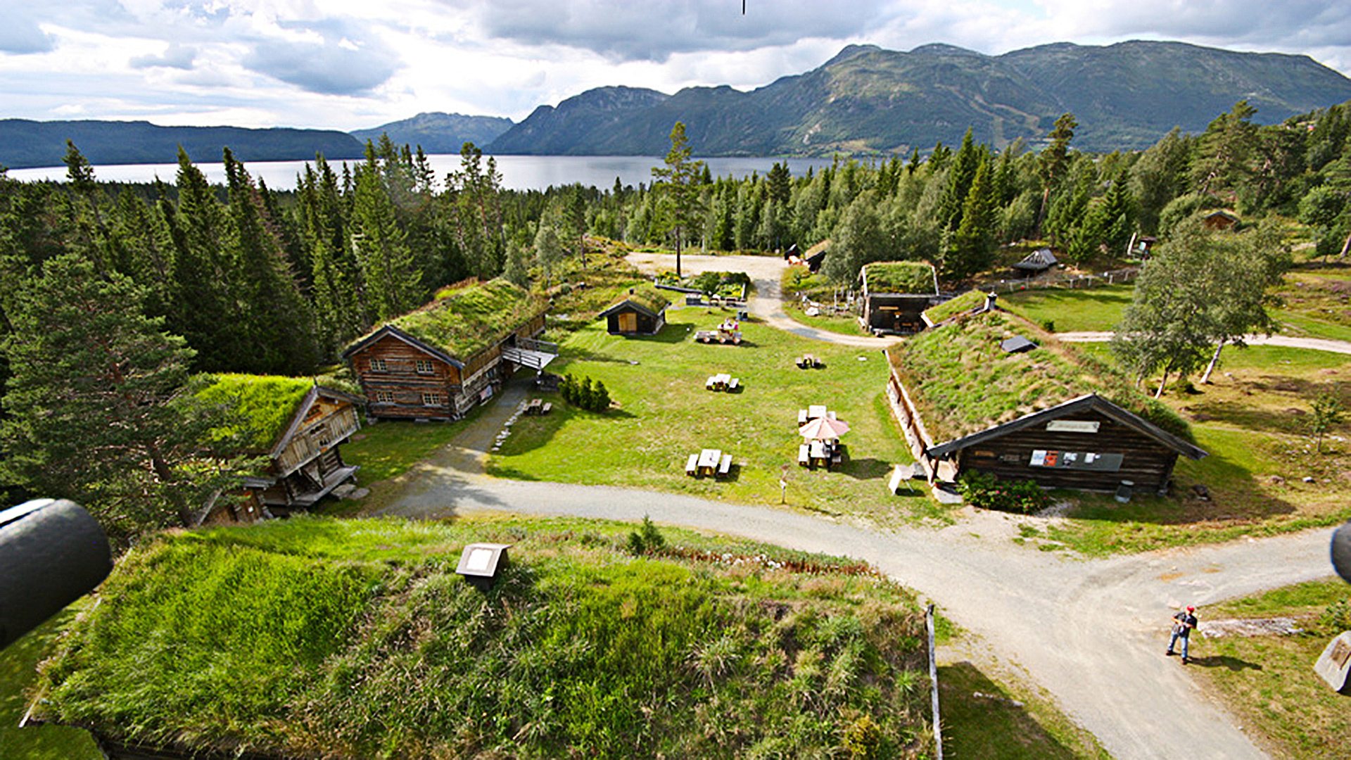 Norsk gårdstun med tradisjonelle torvtakshus omgitt av skog og fjell ved en innsjø.