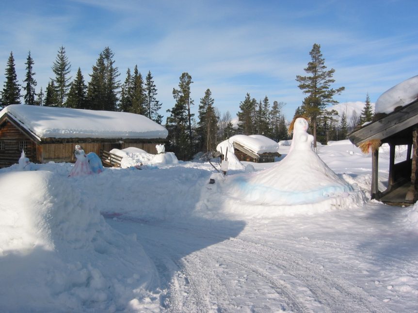 Snøskulpturer av kvinner i en vinterhage med trehus og snødekte omgivelser.