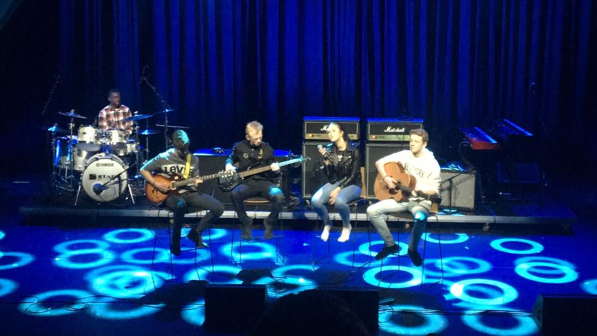 Band spiller på scenen med to gitarister, sanger, trommeslager og opplyste sirkelmønstre på gulvet.