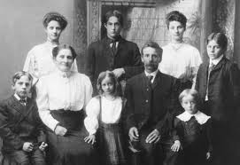 Gammelt familiefoto av ni personer i formelle klær, sittende og stående, med et dekorativt bakteppe.