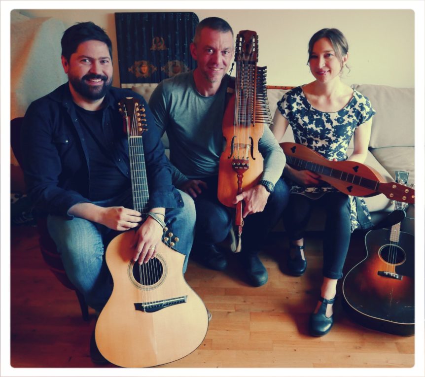 Tre musikere med gitar, nyckelharpa og dulcimer sitter sammen i en stue.