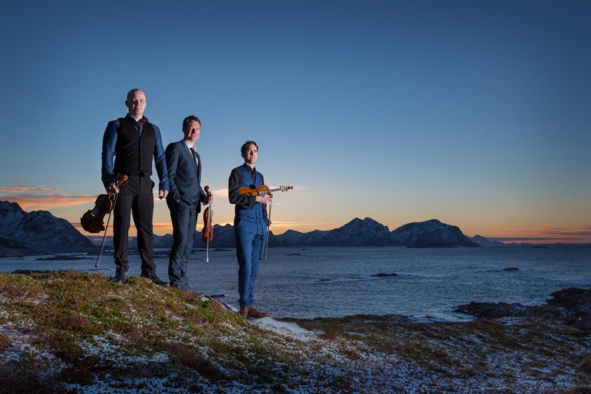 Tre menn med fioliner står på kystklippe ved solnedgang i Norge, i bakgrunnen ses snødekte fjell og hav.
