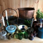 Antikke glass- og keramikksamling på trebord med planter i bakgrunnen.